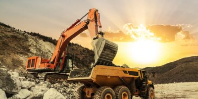 Excavator loading dumper truck on mining site at sunset.